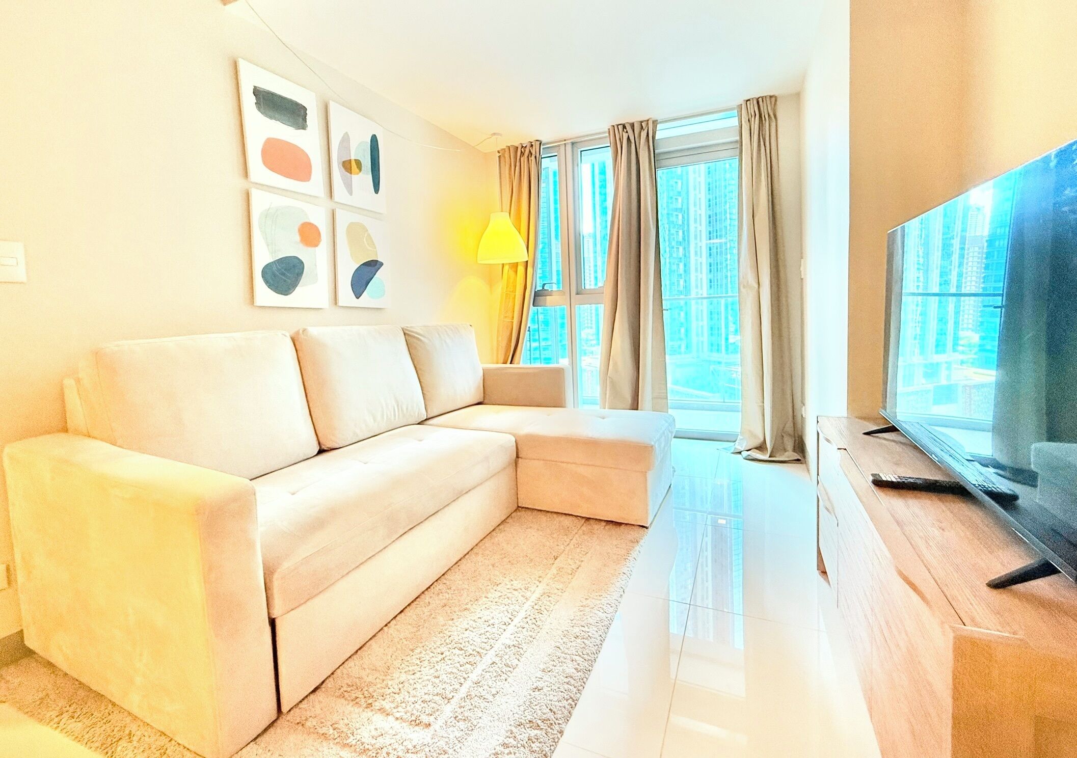 A modern living room with a beige sectional sofa, a flat-screen TV on a wooden stand, and a large window with curtains.