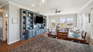 A spacious living room with a large entertainment unit, a dining area with a granite countertop, and a view of the outdoors through large windows. - Surfside Retreat - Great Views & Steps from Beach (Freeport)