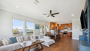 A spacious living area with a sofa, coffee table, and a dining table. The kitchen is visible in the background with wooden cabinets and a TV. - Surfside Retreat - Great Views & Steps from Beach (Freeport)