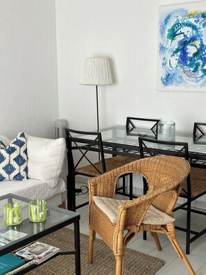 A living room with a sofa, a wicker chair, a glass coffee table, a lamp, and a painting on the wall.