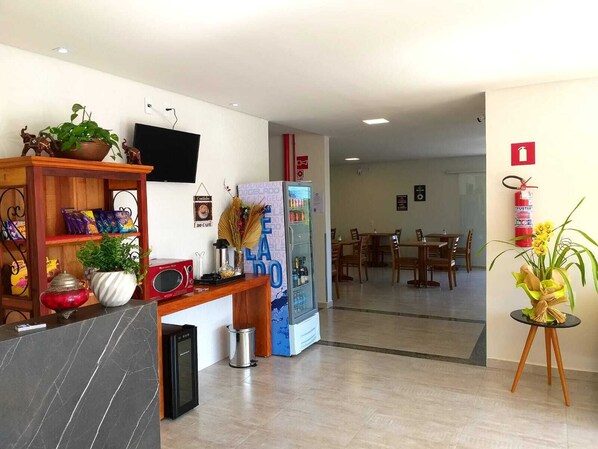 A hotel lobby with a reception desk, a television, a vending machine, and a dining area in the background.