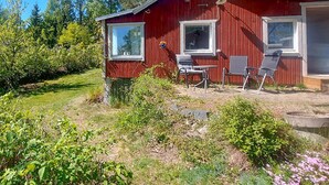 Punainen puutalo, jossa on kaksi ikkunaa, terassi ulkokalusteineen ja puutarha, jossa kasvaa monenlaisia kasveja.