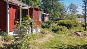 Punainen puutalo, jossa on kuisti, ja sen ympärillä on vehreää kasvillisuutta sekä polku, joka johtaa vesistöön.