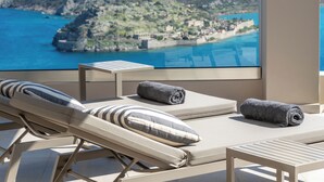 A balcony with a view of a coastal town, two lounge chairs with towels, and a small table. - Notiko villas (Agios Nikolaos)