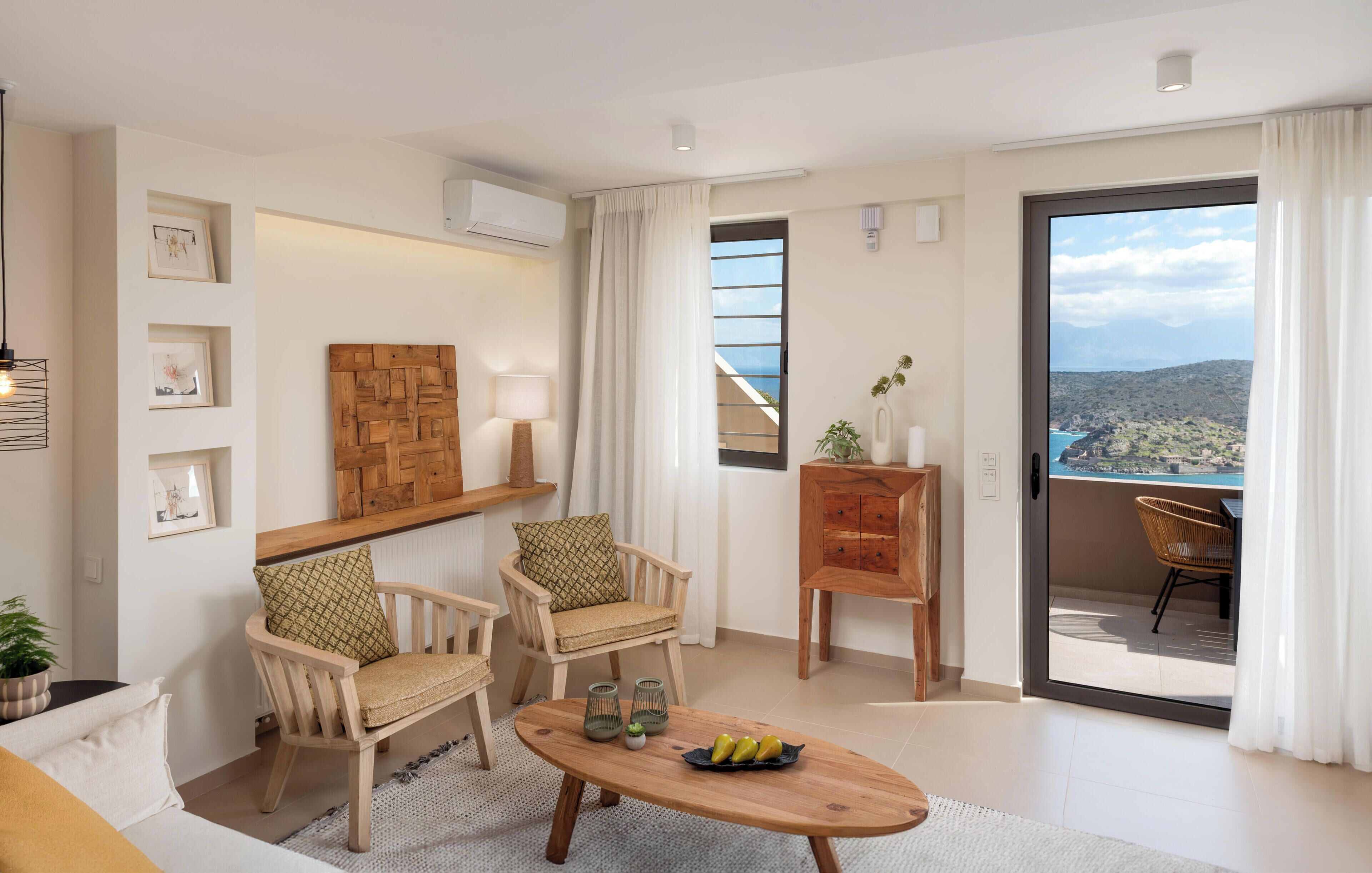 Una sala de estar moderna con dos sillas de madera, una mesa de centro de madera con frutas, una planta en maceta y vistas al mar a través de un ventanal grande.