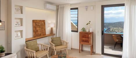 Una sala de estar moderna con dos sillas de madera, una mesa de centro de madera con frutas, una planta en maceta y vistas al mar a través de un ventanal grande.