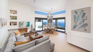 A spacious living room with a gray sofa, a dining table with chairs, and a view of the sea through large windows. - Notiko villas (Agios Nikolaos)