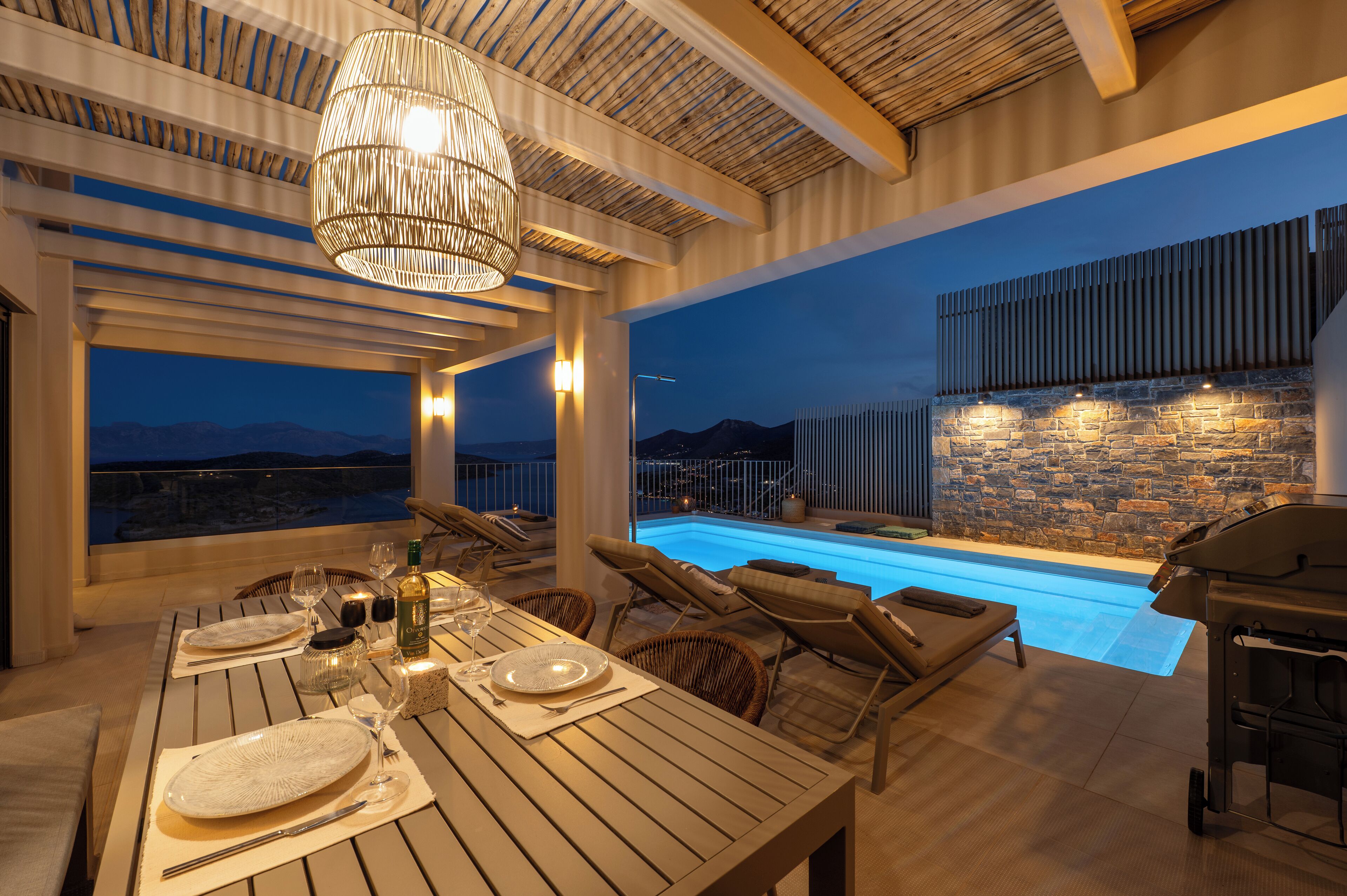 A dining area with a pool view, wooden furniture, and a stone wall.