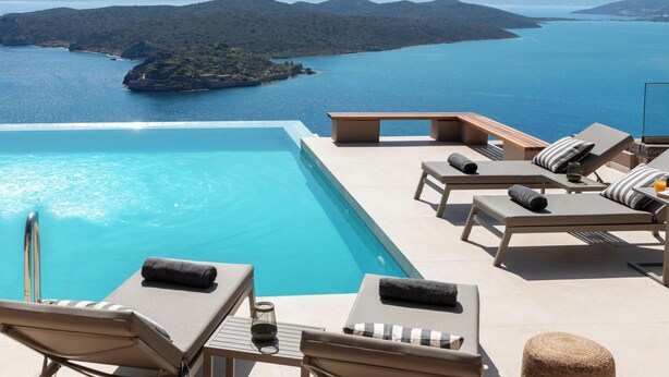An infinity pool with lounge chairs and a view of a large body of water and distant land. - Notiko villas (Agios Nikolaos)