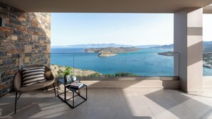 A balcony with a stone wall, a wicker chair, a small table with a plant, and a view of the sea and distant mountains. - Notiko villas (Agios Nikolaos)