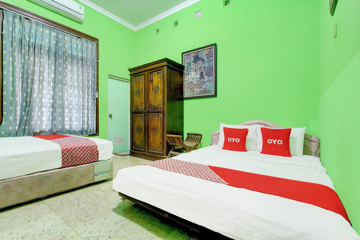 A hotel room with two beds, a wooden wardrobe, a green wall, and a framed artwork.