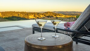 Una terraza con vistas a un paisaje montañoso, dos copas de martini sobre una mesa de mimbre y una silla de lounge con una toalla rosa.