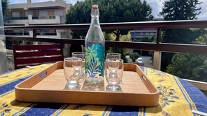 A balcony with a wooden tray holding a bottle and glasses, a patterned tablecloth, and a view of buildings and trees. - Studio de Saint-laurent 200m de la Plage (Saint-Laurent-du-Var)