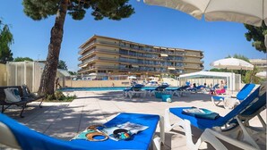 A poolside area with lounge chairs, umbrellas, and a building with multiple balconies in the background. - Studio de Saint-laurent 200m de la Plage (Saint-Laurent-du-Var)