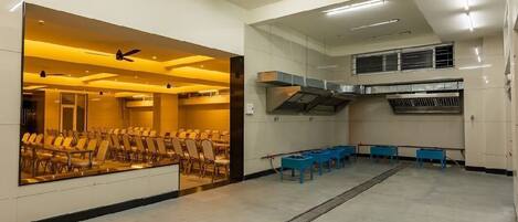 Sala de conferência com fileiras de cadeiras, espaço de cozinha e ventiladores no teto.