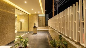 A modern hotel entrance with a glass door, a reception desk, and a row of potted plants along the walkway. - Patel Comforts Inn & Suites (Mysore)