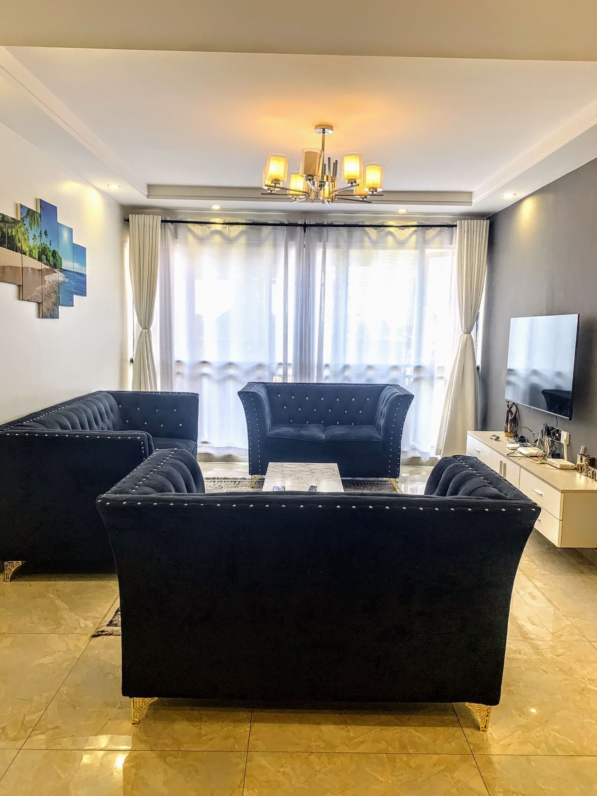 A modern living room with a dark blue sofa set, a chandelier, a flat-screen TV, and a large window with curtains.