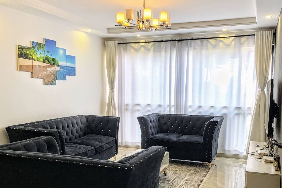 Una sala de estar moderna con un sofá negro, una lámpara de araña y un ventanal con cortinas.