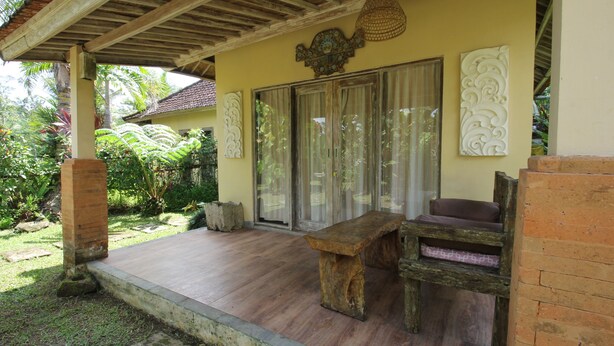 A covered outdoor seating area with a bench and a cushioned chair, a wooden table, and decorative wall art. - Capital O 94012 Dewa Jati House (Sebatu)