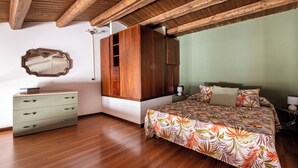 Ein Schlafzimmer mit Holzboden, einem Bett mit Blumenmuster, einem Kleiderschrank, einer Kommode und einer Decke mit sichtbaren Holzbalken.