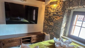 A hotel room with a stone wall, a flat-screen TV mounted above a wooden console table, and a bed with a green bedspread and two towels shaped like flowers. - Old house Njivice (Omisalj)