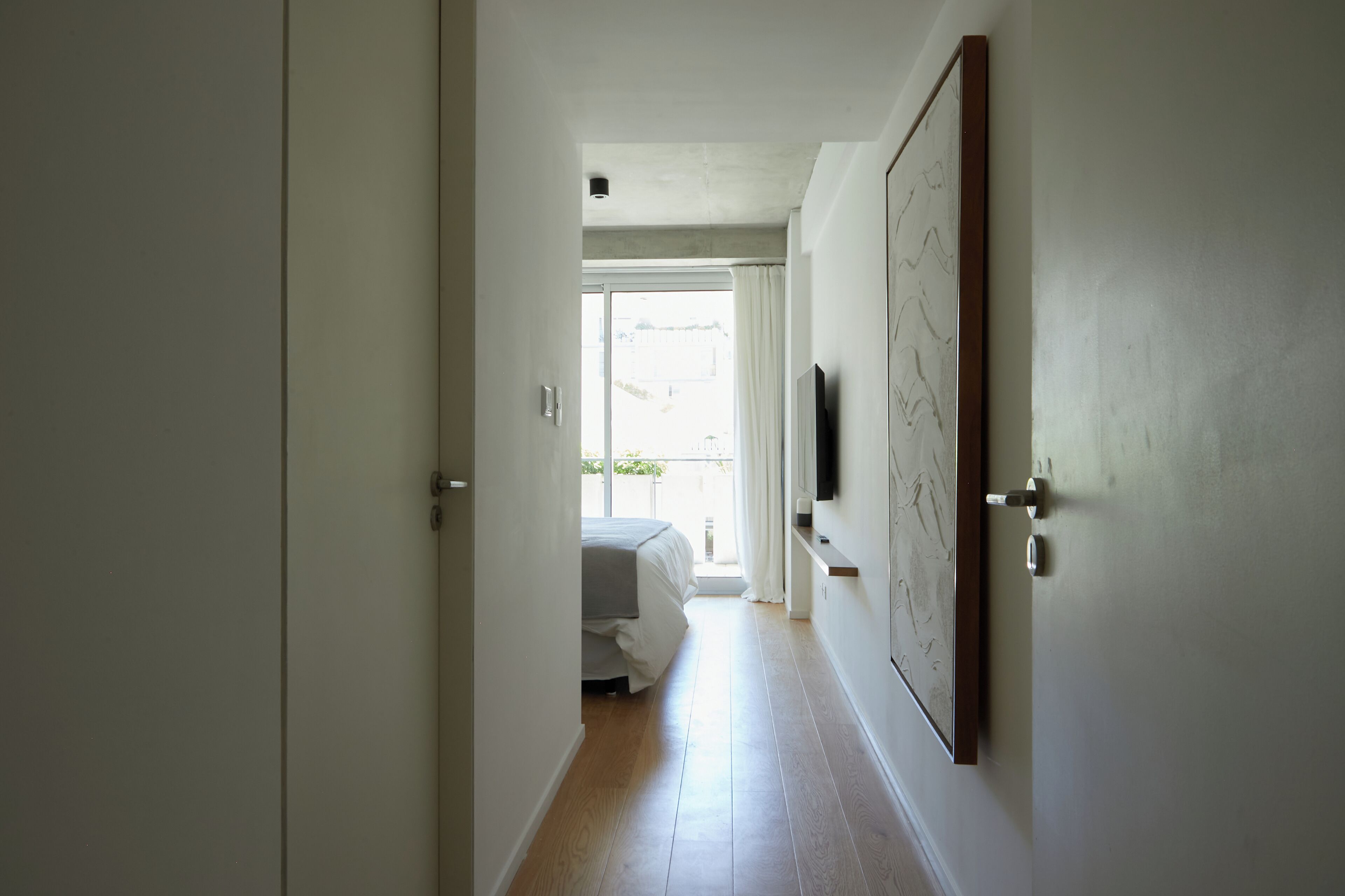 A narrow hallway with a bed at the end, a door on the right, and a window with curtains.
