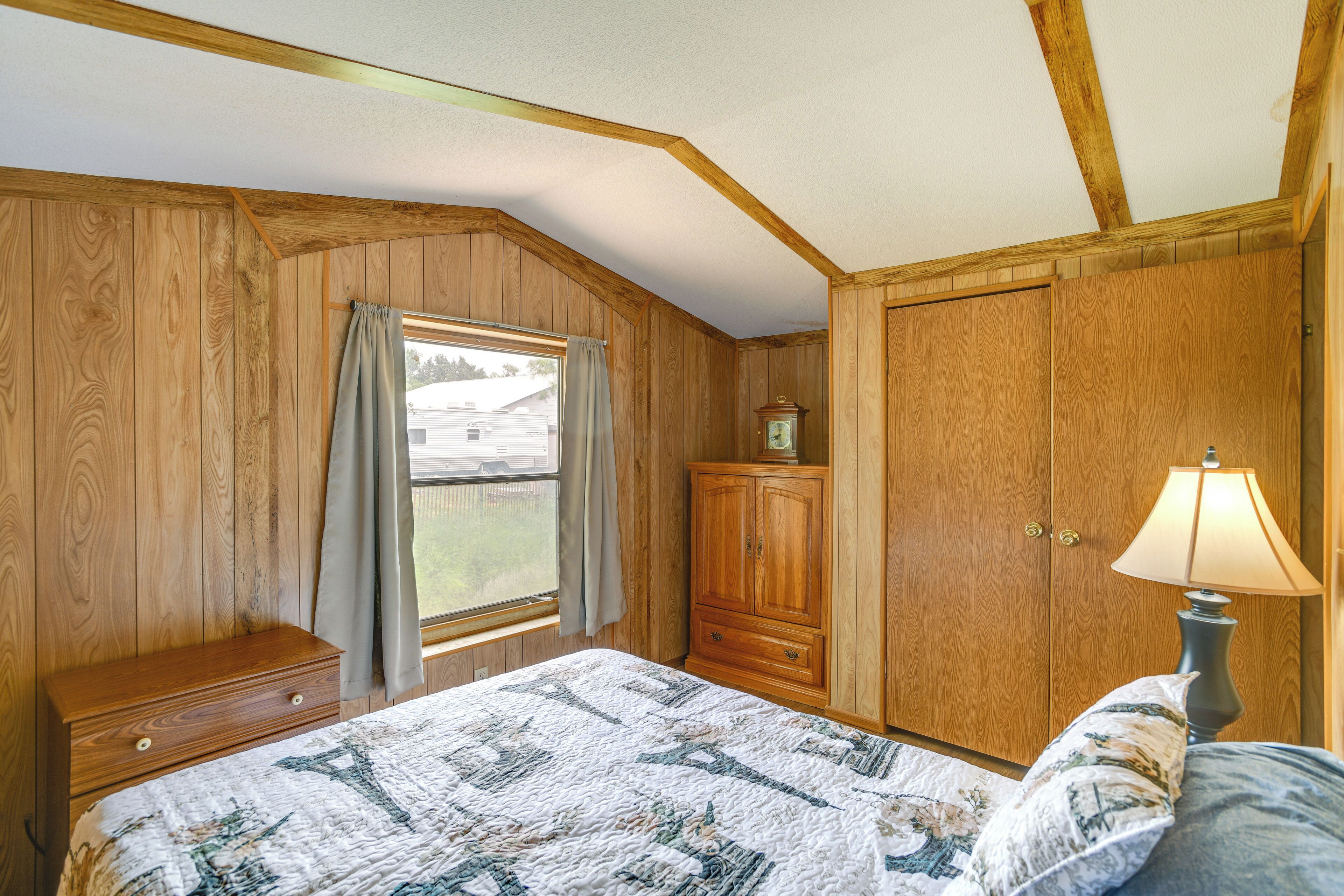 A bedroom with wooden walls, a bed with a quilt, a nightstand, a lamp, and a window with curtains.