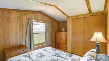 A bedroom with wooden walls, a bed with a quilt, a nightstand, a lamp, and a window with curtains.