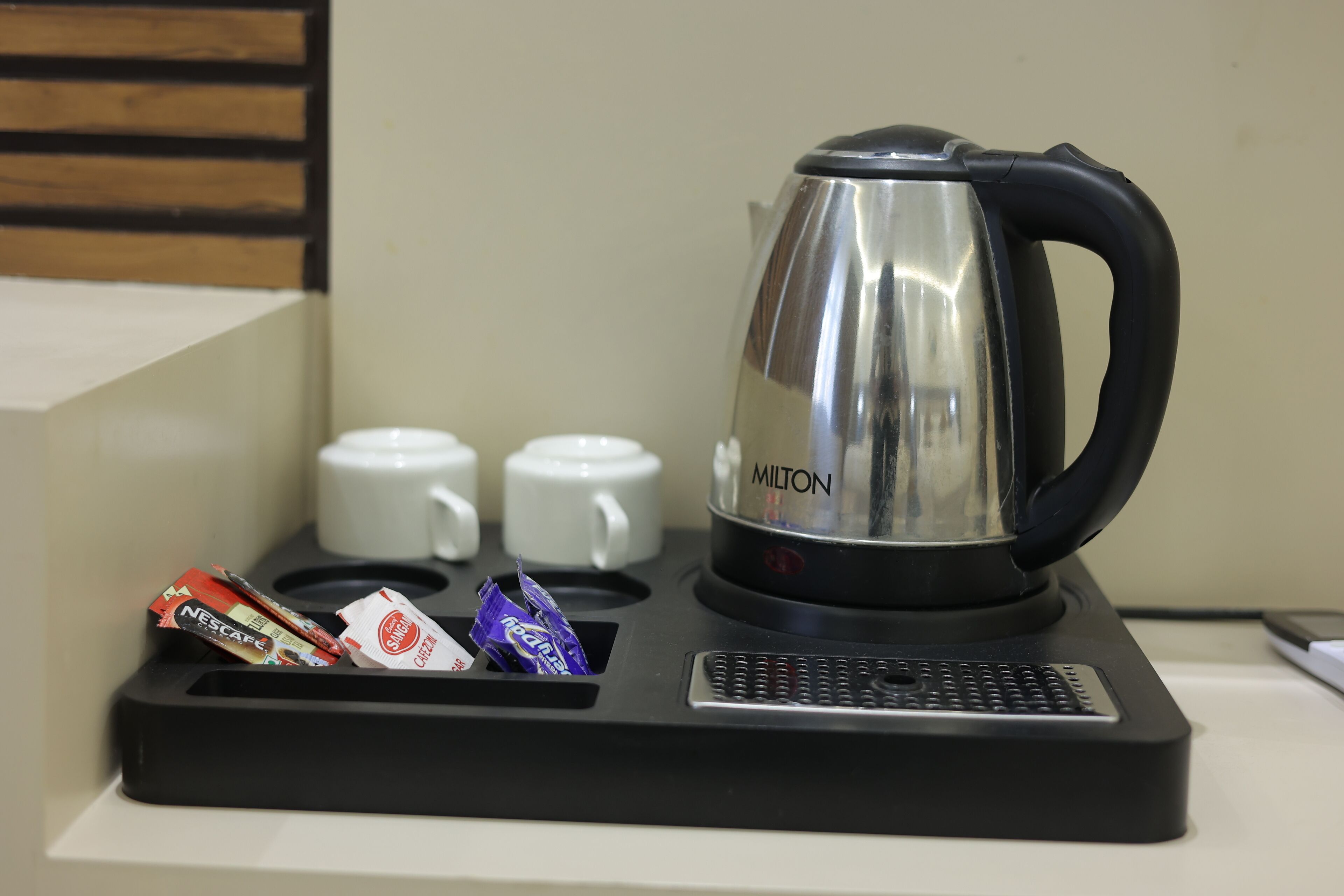 Una macchina per il caffè con manico nero e un bollitore in acciaio inossidabile con la scritta "MILTON" su un vassoio nero, con due tazze bianche e un portacapsule.