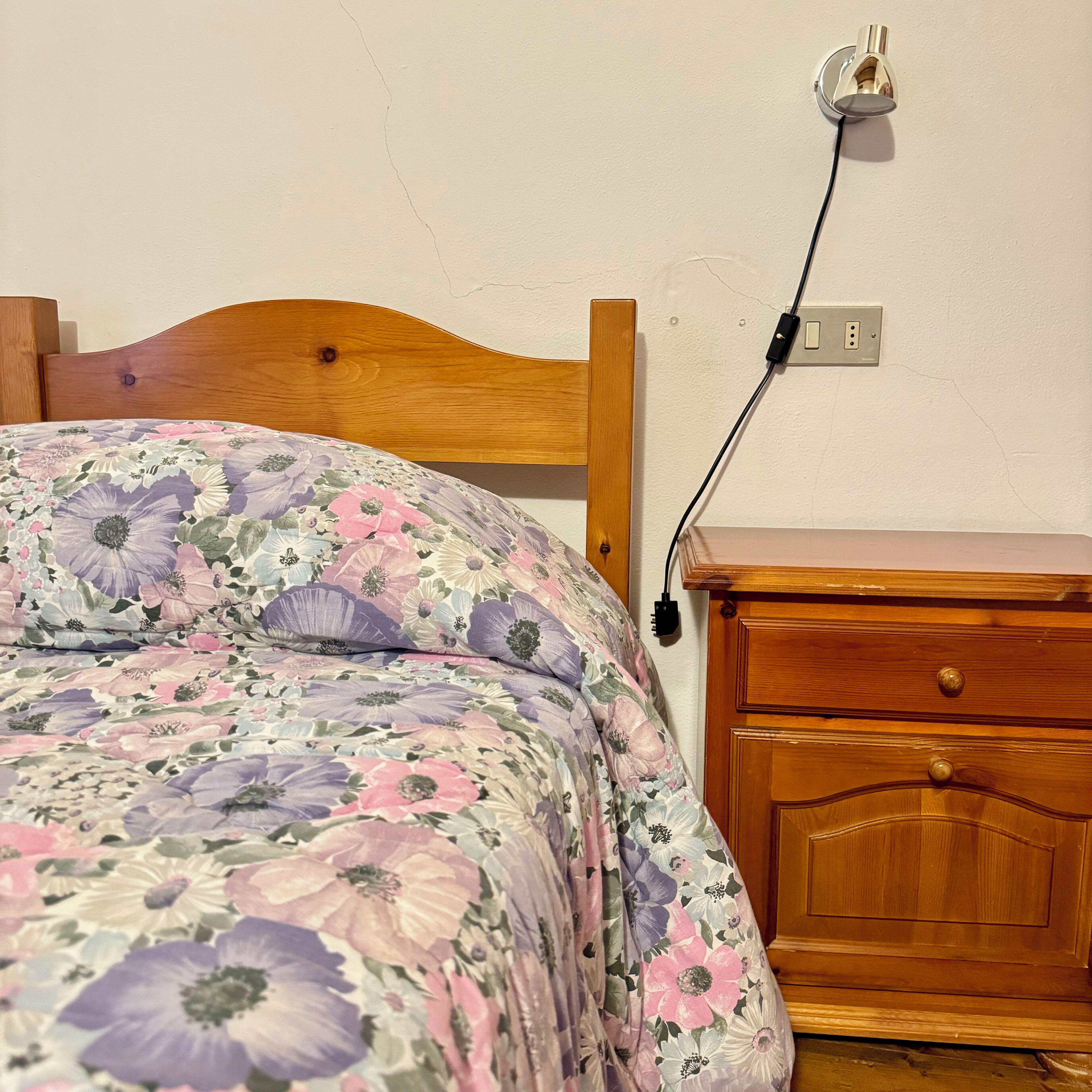 Een bed met een bloemenprintdeken, een houten hoofdbord, een nachtkastje met lades, en een stopcontact.