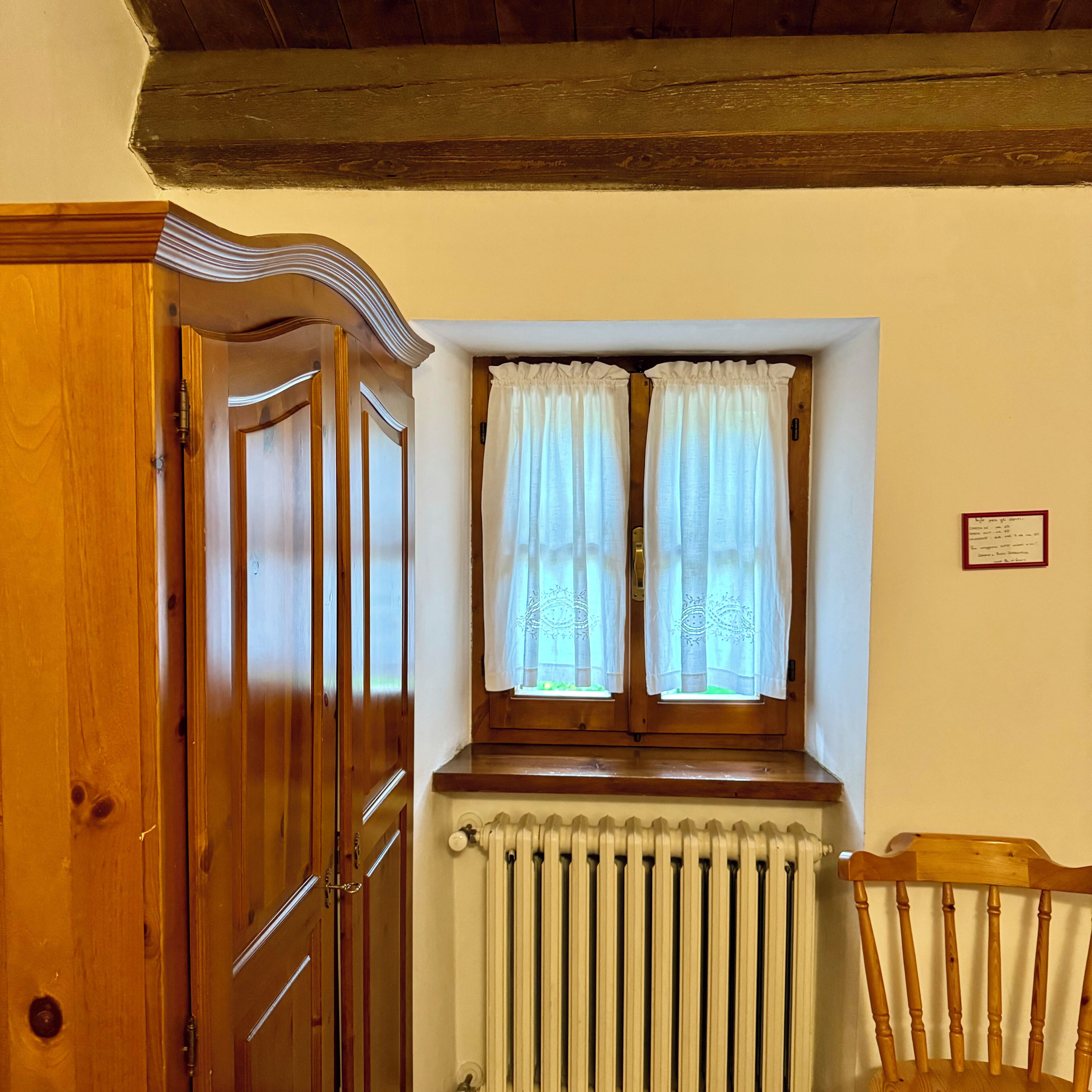 A room with a wooden door, a window with white curtains, a radiator, and a wooden chair.