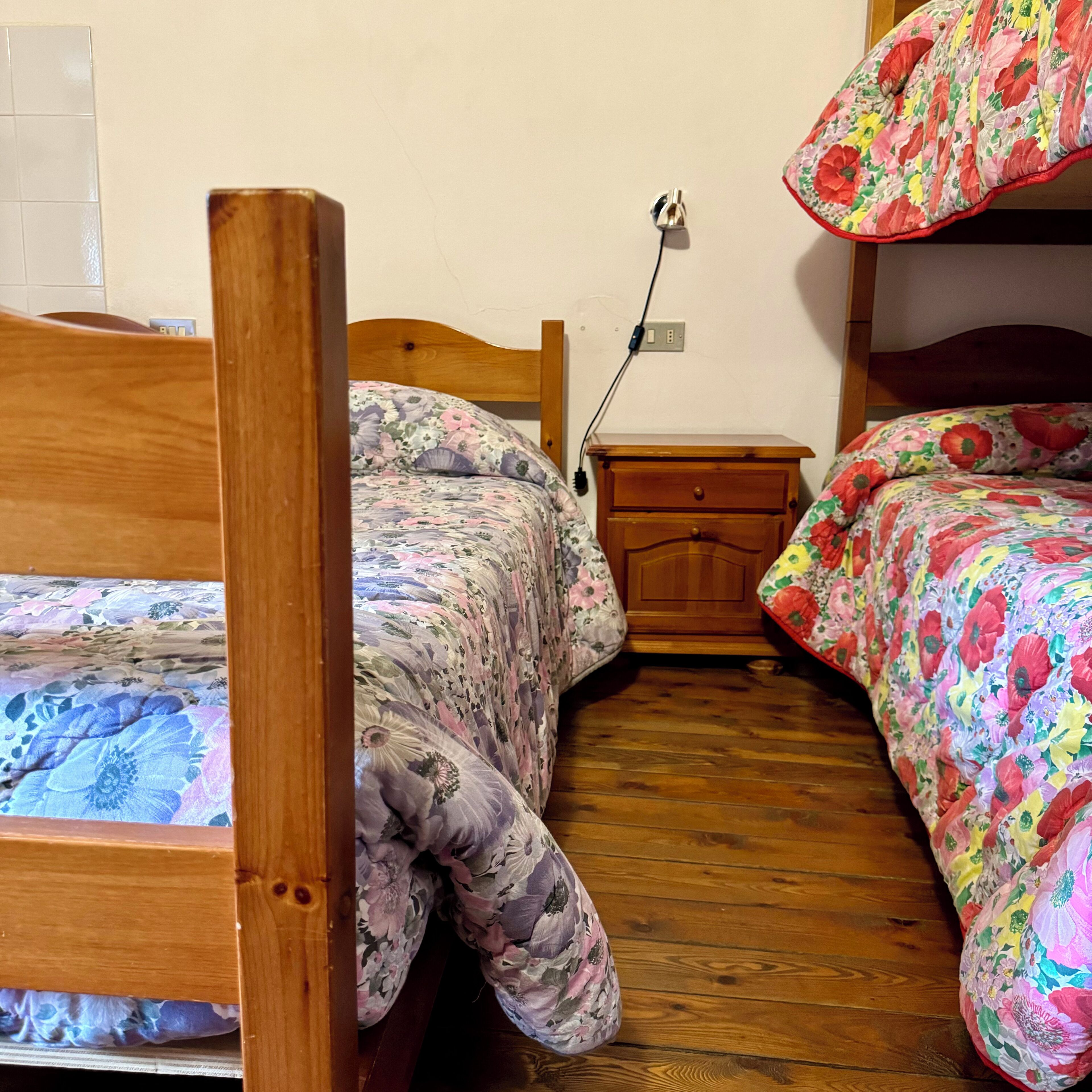 Une chambre à coucher avec deux lits en bois, chacun agrémenté de draps à motifs floraux. Une table de chevet en bois, dotée d’un tiroir, se trouve entre les deux lits. Une prise de courant est visible sur le mur.