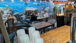 En utomhusrestaurang med stenmurar, ljusslingor och en väggmålning. Bord och stolar är uppställda för att skapa en trevlig matplats.