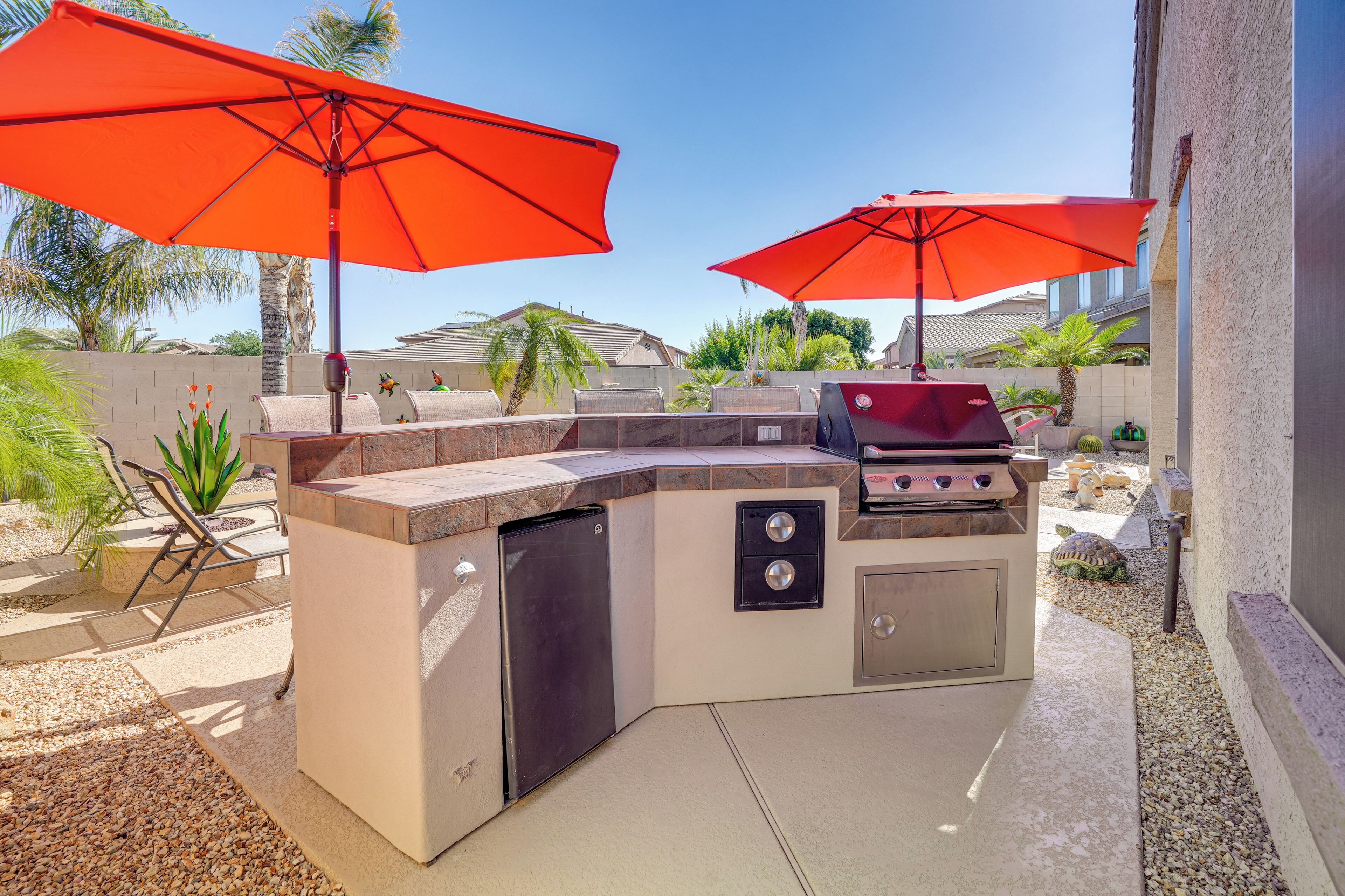 An outdoor kitchen with two red umbrellas, a grill, and a built-in refrigerator.