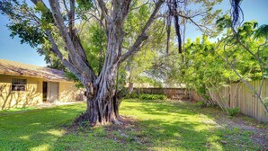 A backyard with a large tree, a wooden fence, and a house with a yellow exterior. - 7 Mi to Satellite Beach: Melbourne Home w/ Yard (Melbourne)