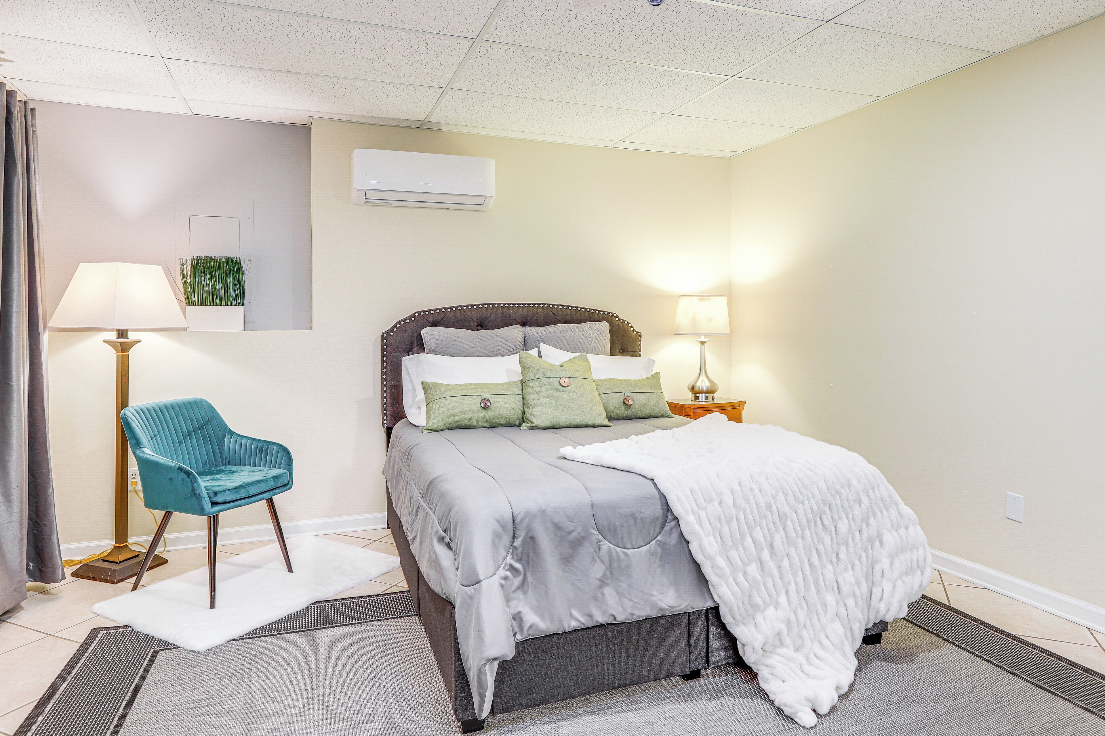 A neatly arranged bedroom with a bed, a chair, a bedside table with a lamp, and an air conditioning unit on the wall.
