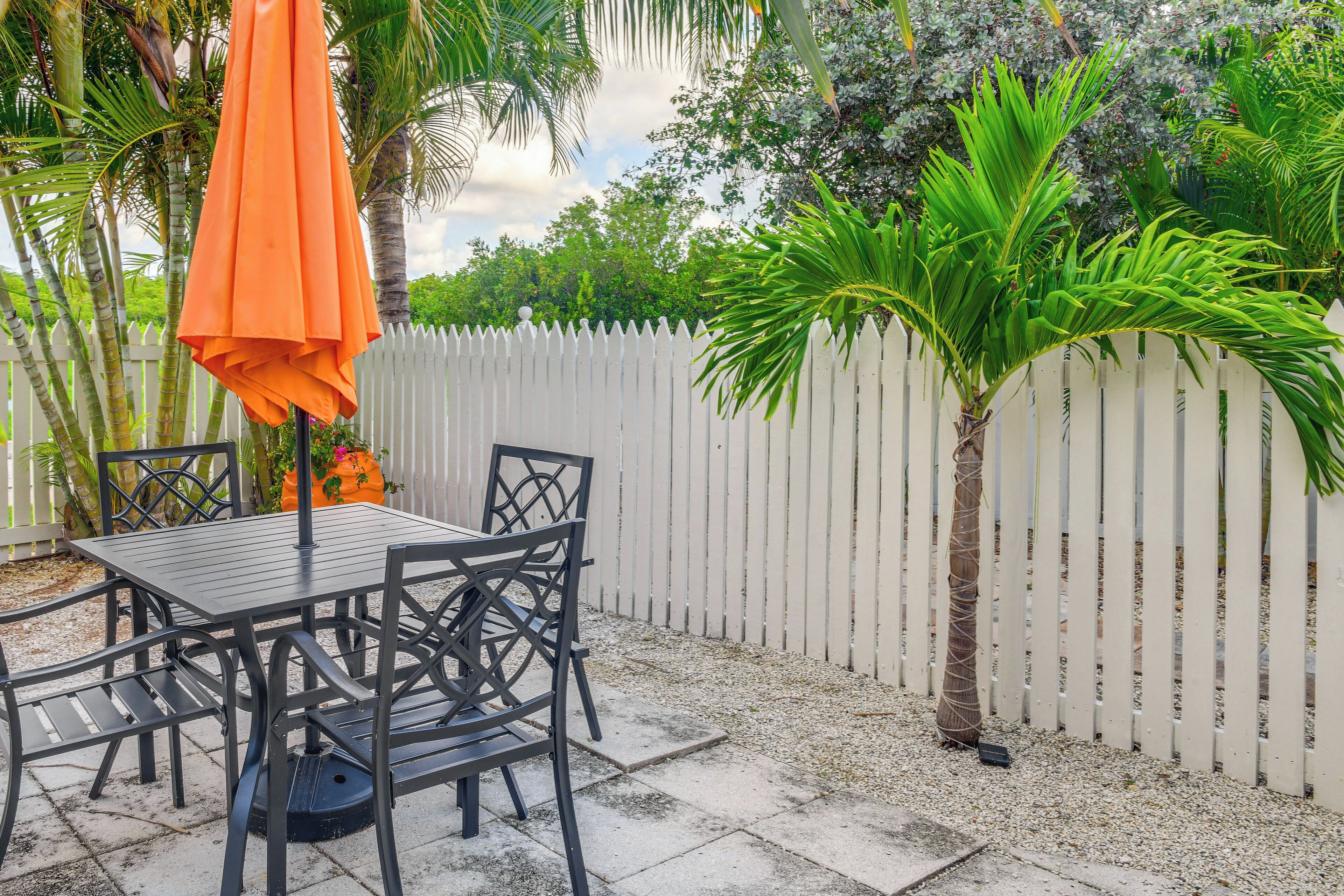 A patio area with a table and chairs, a white picket fence, and tropical plants.