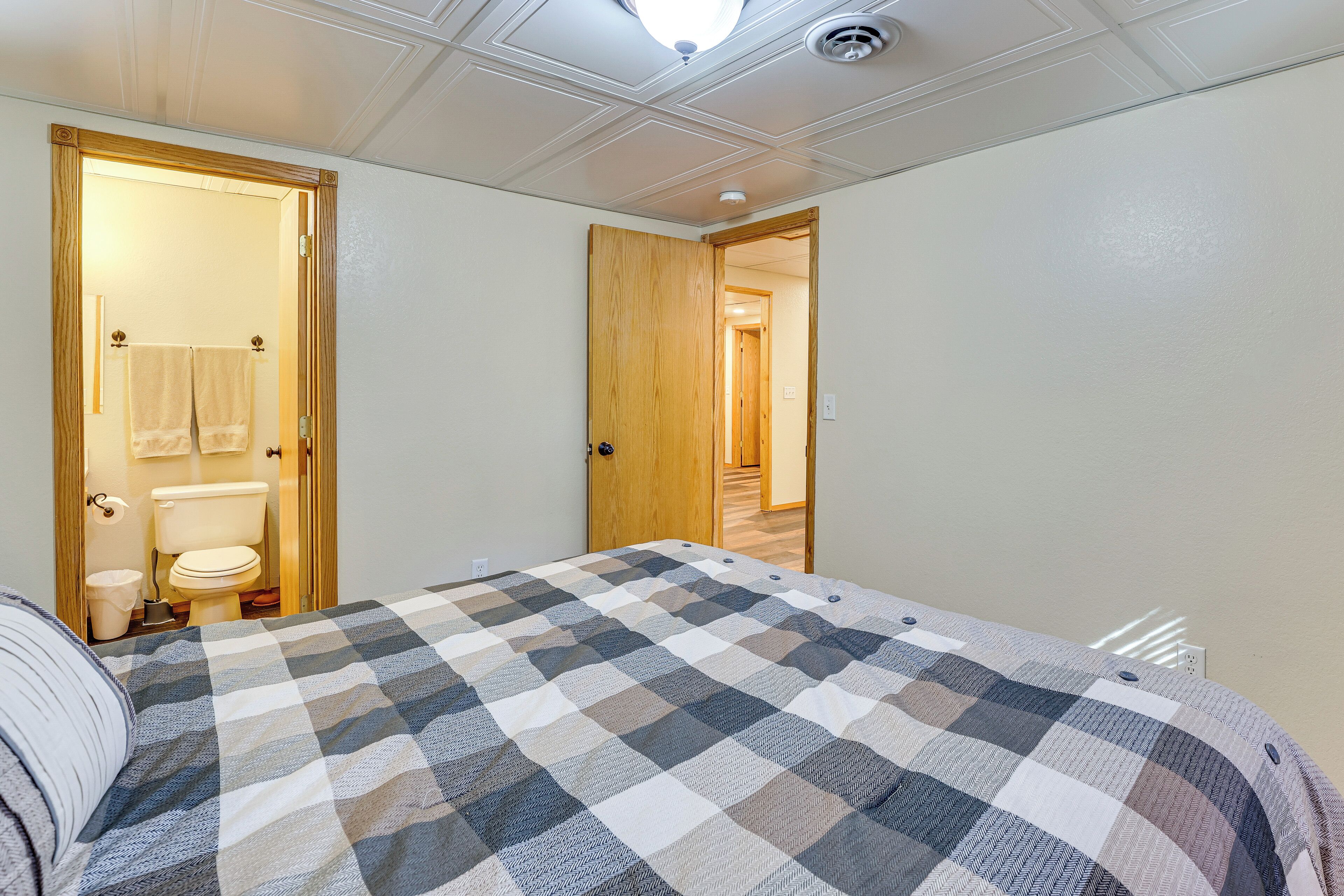 A bedroom with a checkered bedspread, a wooden door leading to a bathroom, and a hallway.