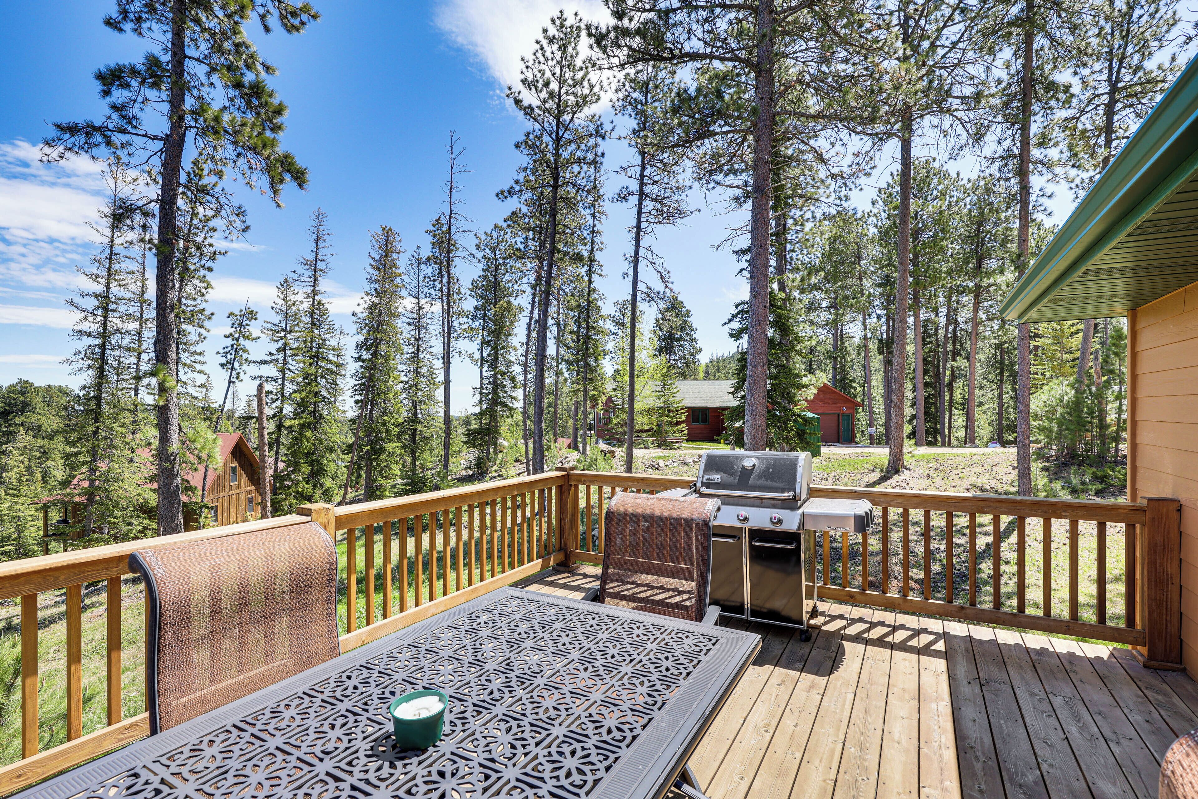 Une terrasse en bois avec un barbecue et un espace salon extérieur, entourée de grands pins.