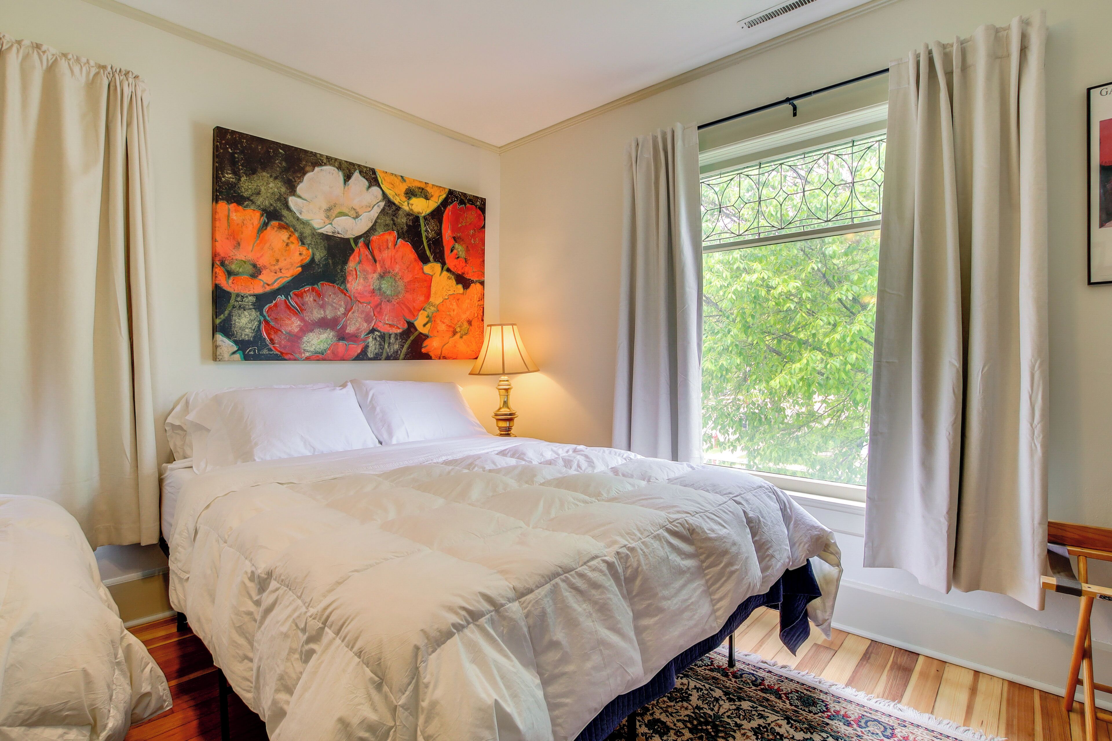 Ein Schlafzimmer mit einem großen Bett, einem Nachttisch, einer Lampe, einem Fenster mit Vorhängen und einem Blumenbild an der Wand.