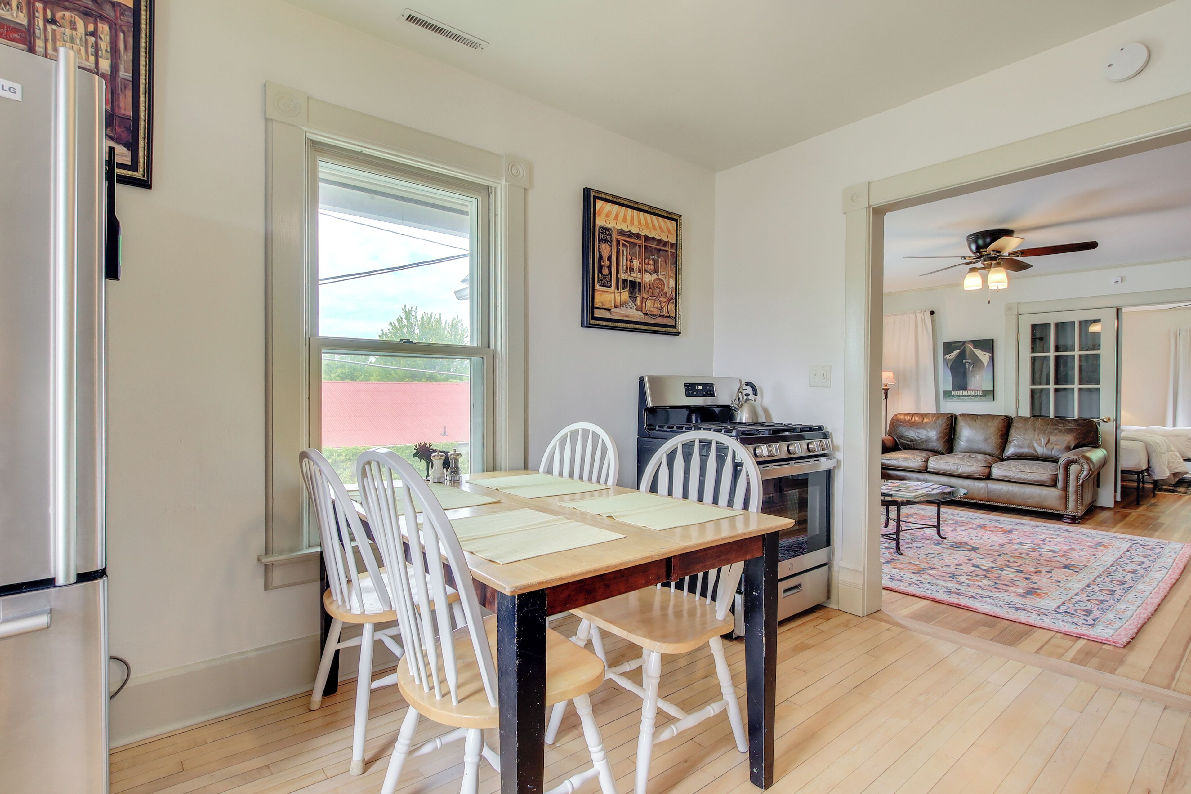 A dining area with a wooden table and white chairs, a kitchen with a stove and refrigerator, a living room with a sofa and a ceiling fan, and a large window with blinds.