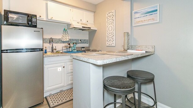 A compact kitchen with white cabinets, a built-in microwave, a stainless steel refrigerator, and a small island with stools.