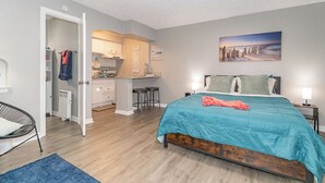 A bedroom with a bed, a chair, a desk, and a kitchen area.