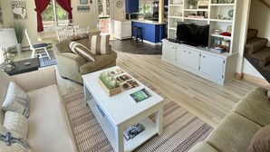 A spacious living room with a sofa set, a coffee table, and a TV. There is a kitchen area with blue cabinets and a dining area with a table and chairs. - Daybreak Limit 8 3 Bedroom Villa by RedAwning (Sebago)