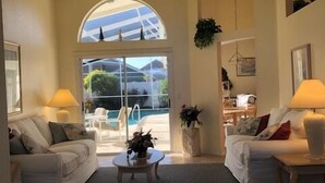 Un salon spacieux avec deux canapés blancs, une table basse et une vue sur une piscine, visible à travers une grande fenêtre en arc de cercle.