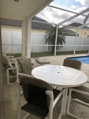 Un patio couvert avec une table ronde et des chaises, une piscine et, au fond, une clôture blanche.
