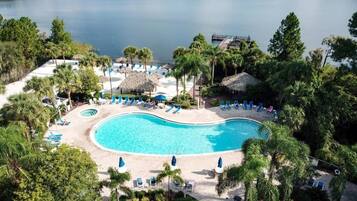 A resort with a large swimming pool, lounge chairs, and a dock extending into a lake.