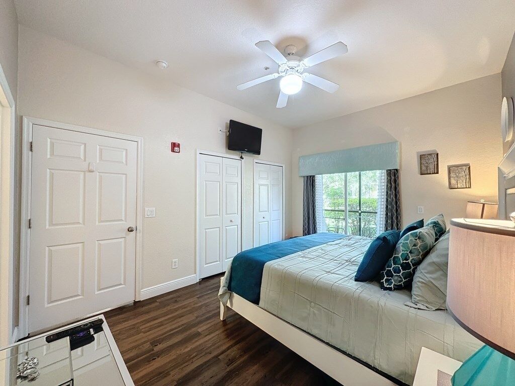 Un dormitorio con cama, ventilador de techo, televisor y ventana con cortinas.