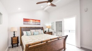A bedroom with a wooden bed, a ceiling fan, a nightstand with a lamp, a painting on the wall, and a door leading to another room.