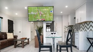 A modern living room with a TV displaying a football game, a dining area with a glass table and chairs, and a kitchen with white cabinets and a herringbone backsplash.